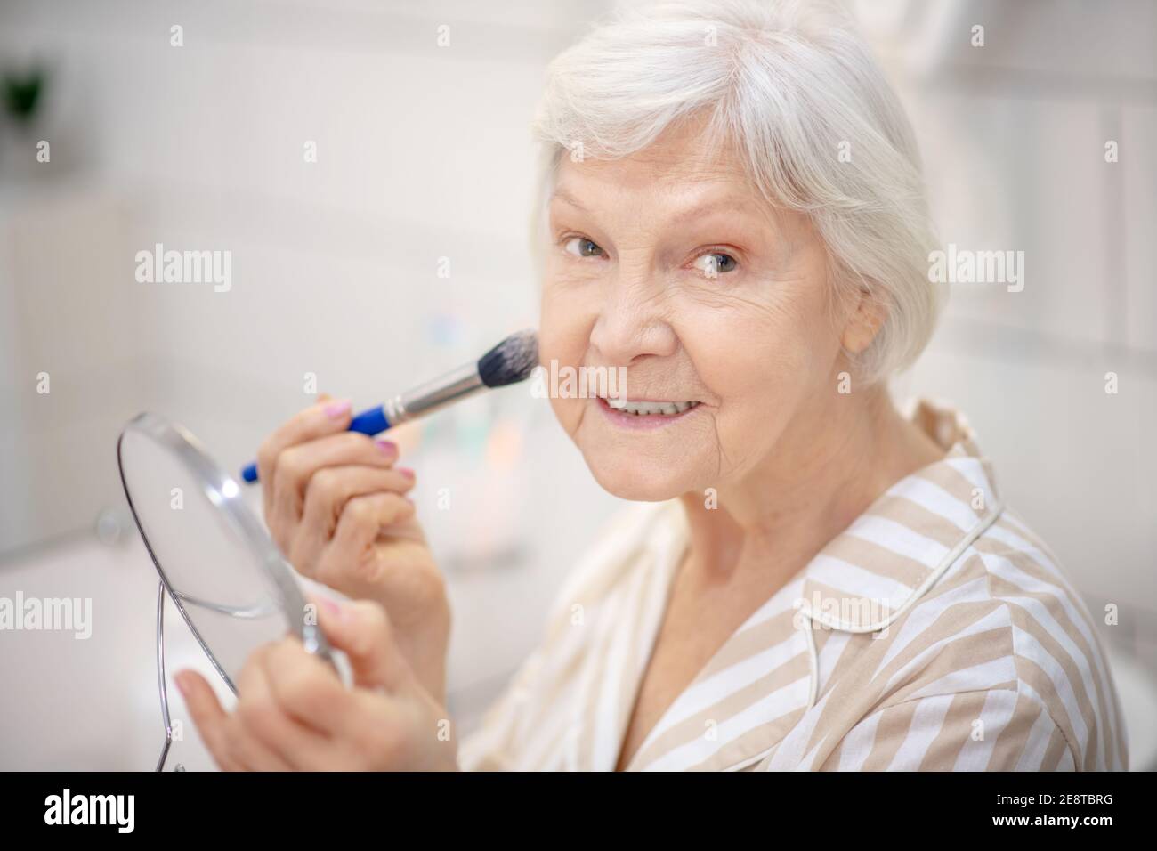 Gray-haired woman in pajama doing make up in the bathroom Stock Photo