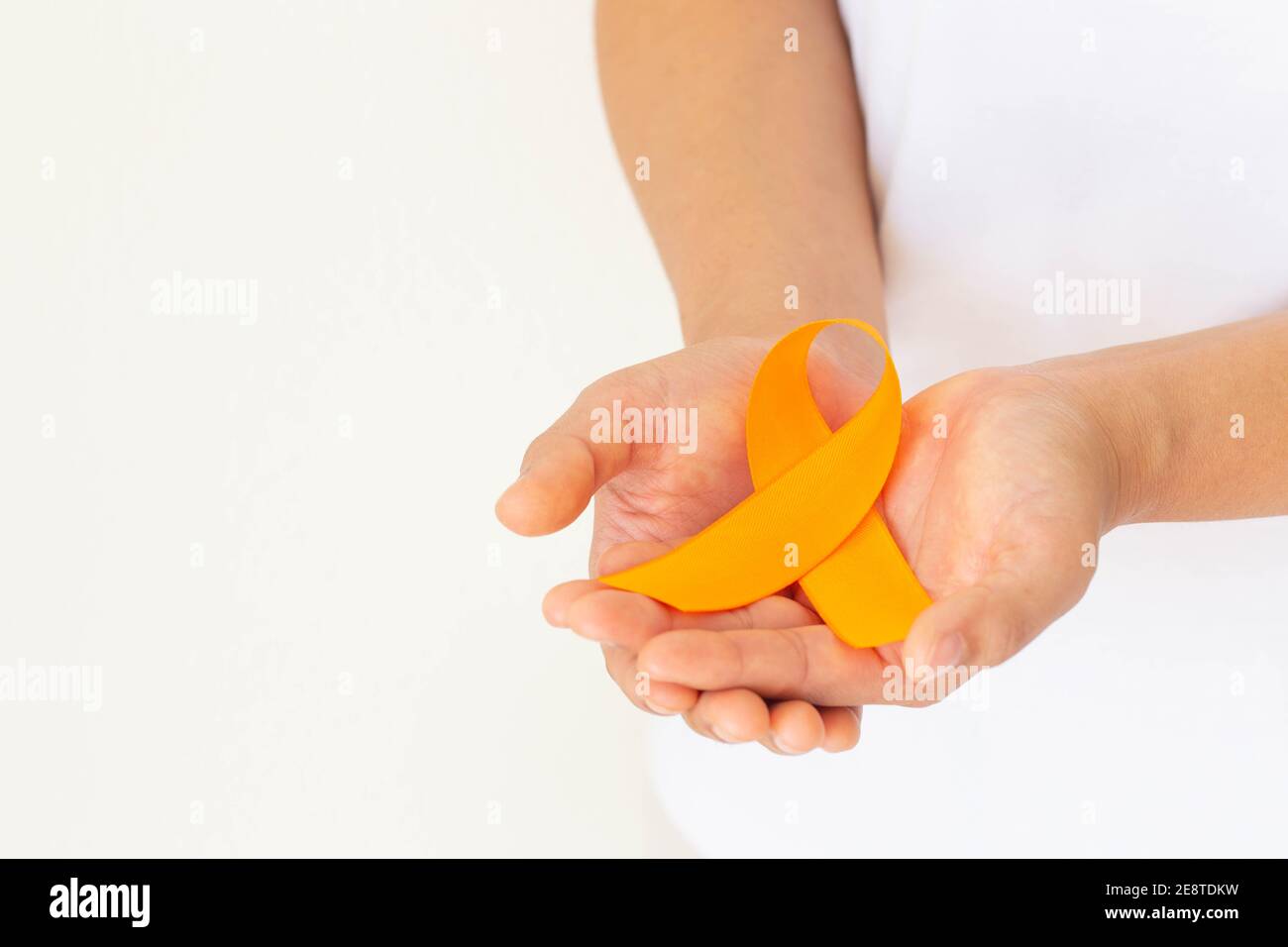 Hands holding orange color ribbon on white fabric with copy space. Kidney Cancer Awareness, Leukemia disease, Skin cancer awareness, World Cancer Day. Stock Photo