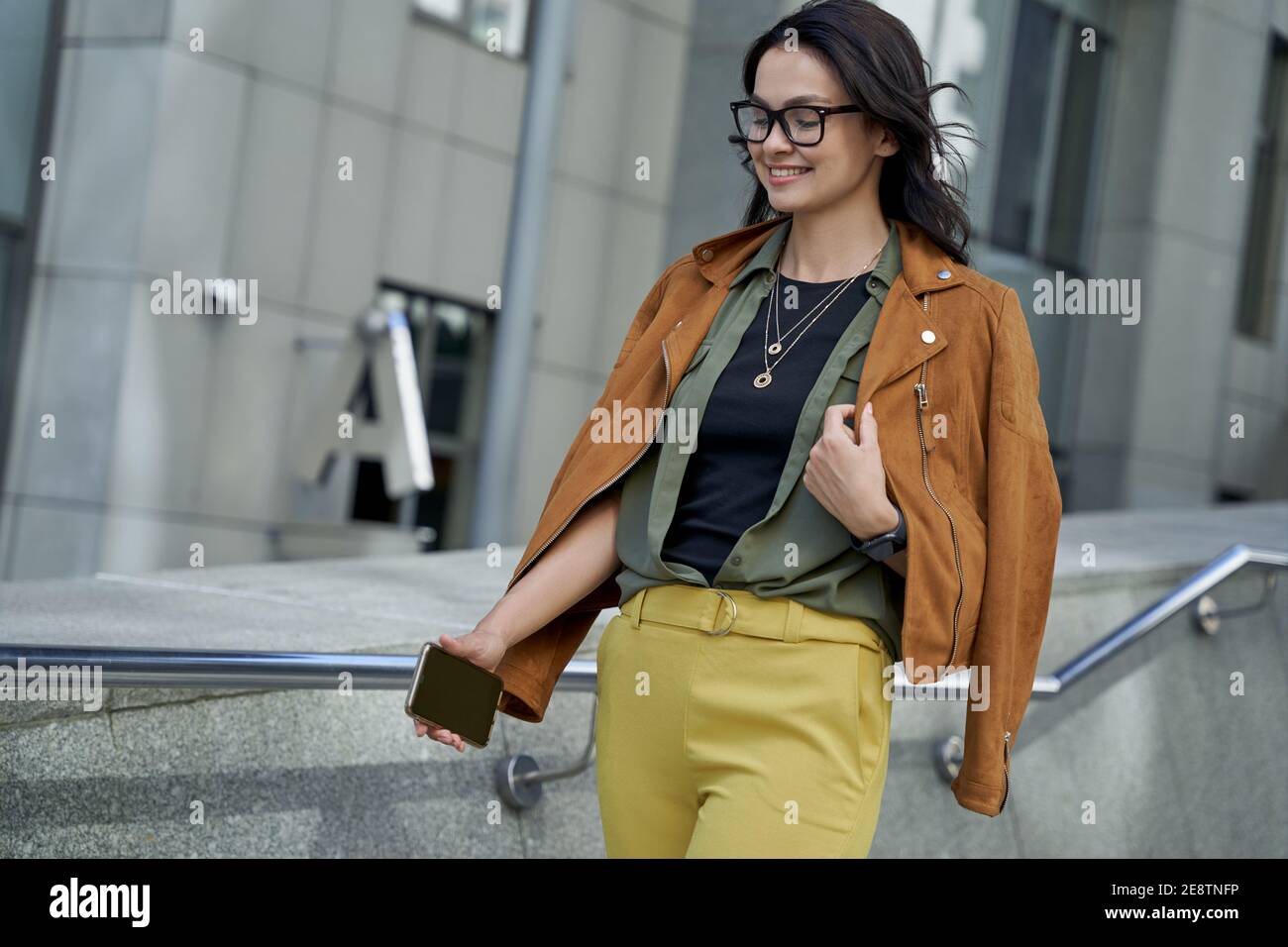 Fashionable girl outdoors. Young beautiful and stylish woman with smartphone in hand walking on the city street and smiling. Urban lifestyle concept, people Stock Photo
