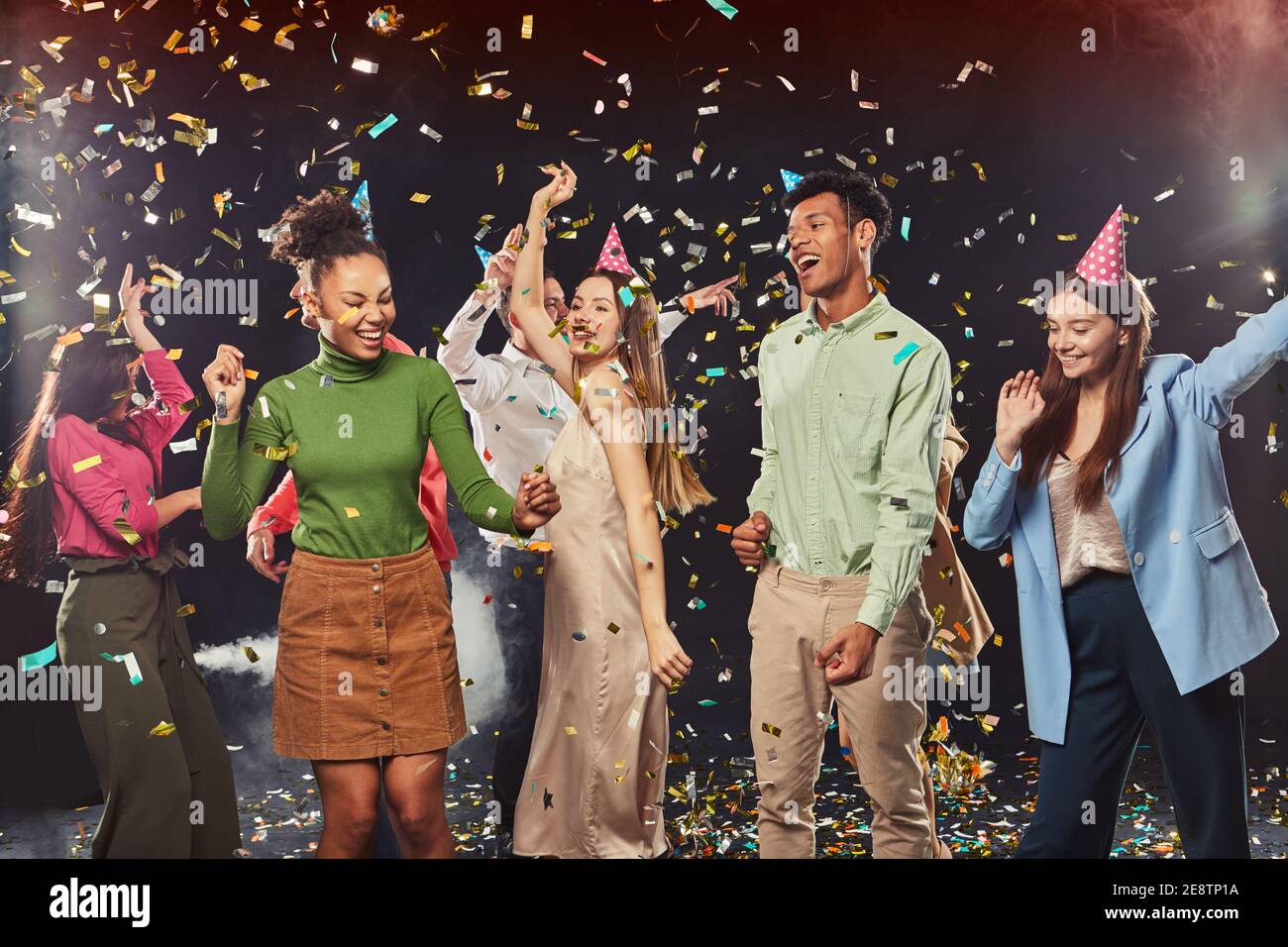 Celebration and party. Group of young happy multiracial people wearing birthday hats dancing and having fun, confetti falling in the air Stock Photo