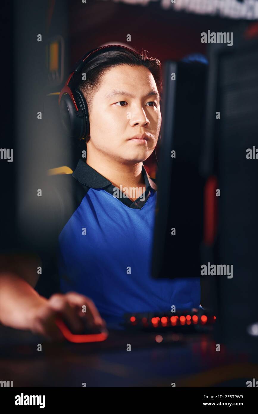 Playing online video games as professional. Vertical shot of concentrated asian guy, male cyber sport gamer wearing headphones looking at PC screen while participating in eSports tournament Stock Photo