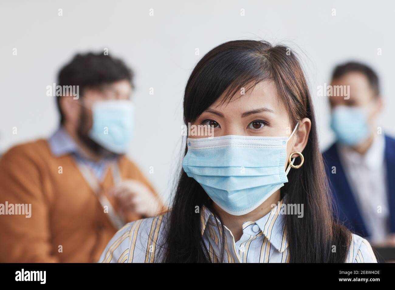 Front view portrait of Asian businesswoman wearing mask and looking at camera with people in background, copy space Stock Photo