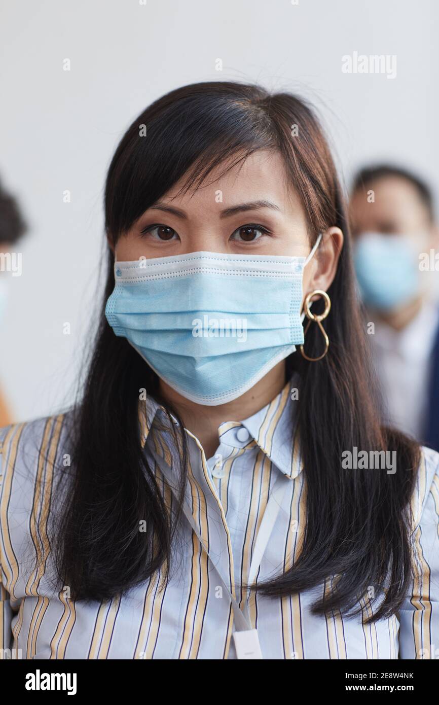 Vertical front view portrait of Asian businesswoman wearing mask and looking at camera with people in background Stock Photo