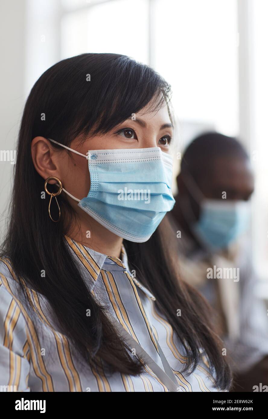 Vertical portrait of Asian businesswoman wearing mask and looking away while sitting in audience at business conference Stock Photo