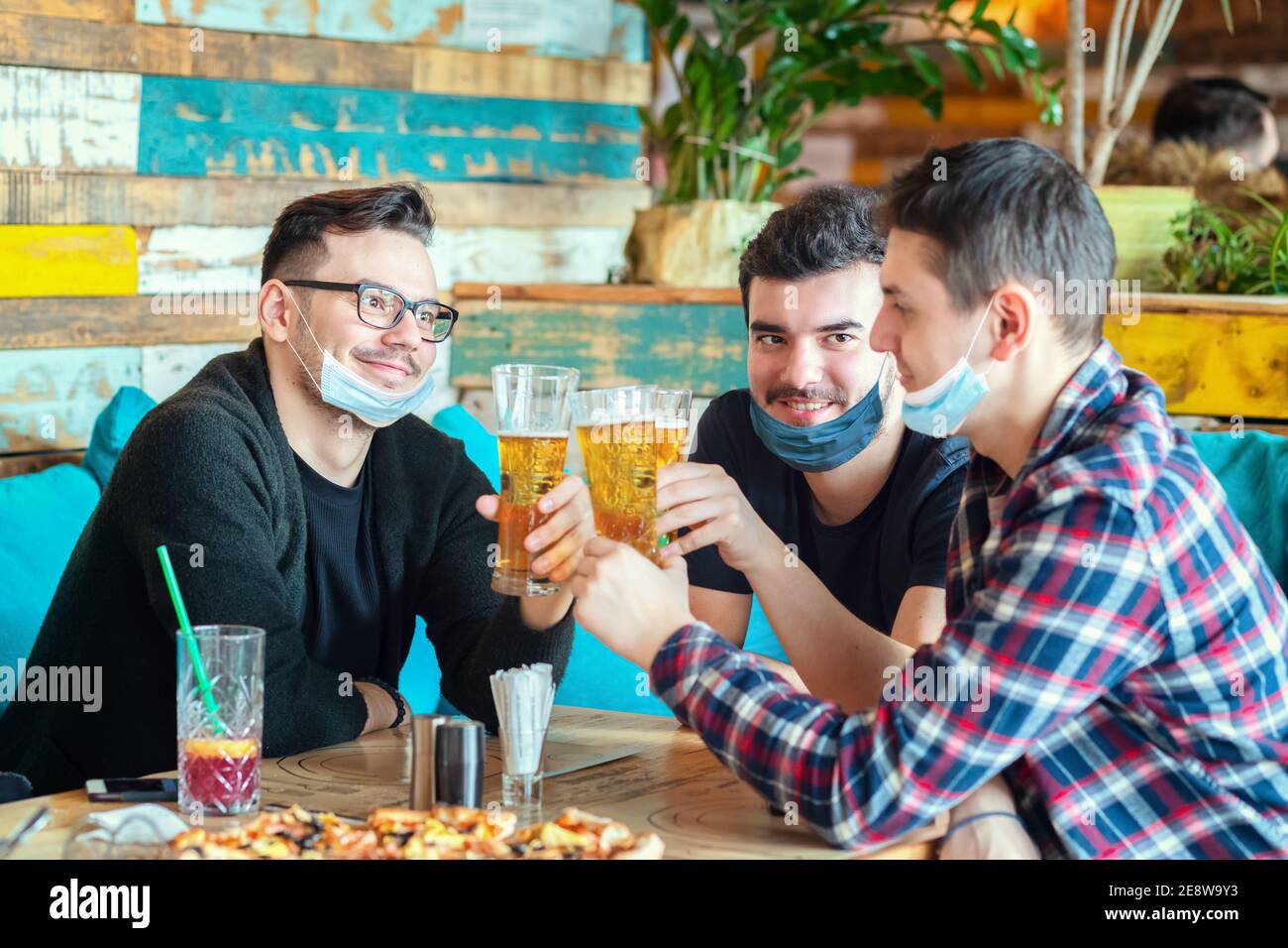 Happy young friends with face mask drinking beer at restaurant table after reopening Stock Photo