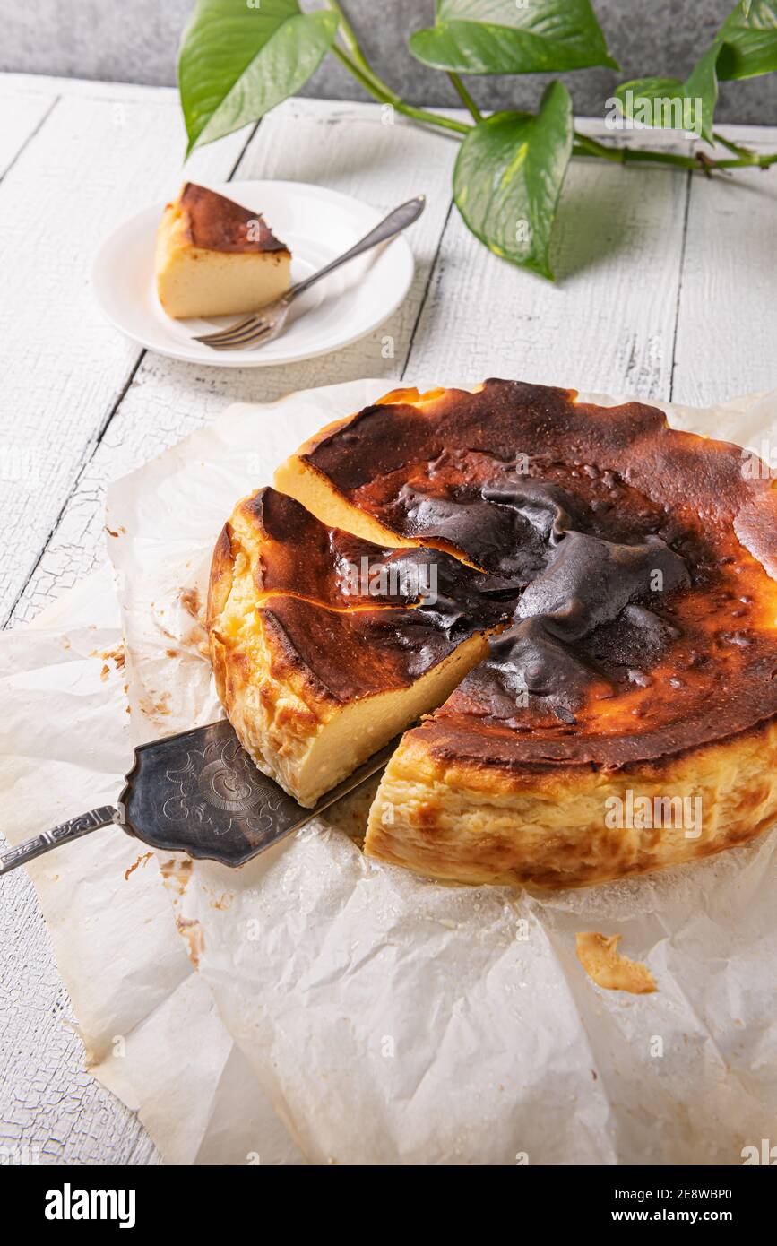 Cut basque burnt cheesecake on silver serving spatula in baking paper on textured white wooden table. Unfocused portion on plate and home plant at bac Stock Photo