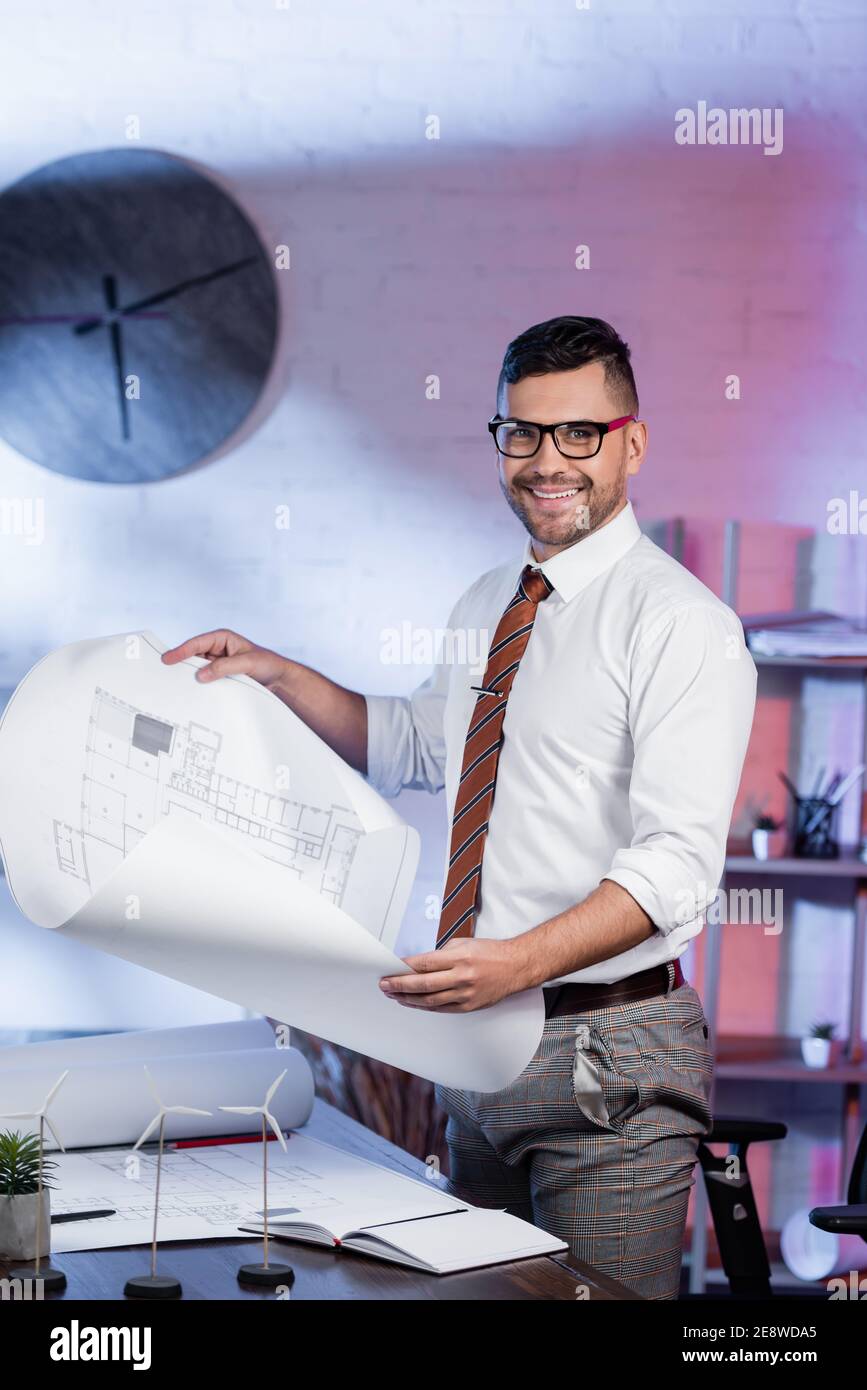 happy architect looking at camera while holding blueprint near models ...