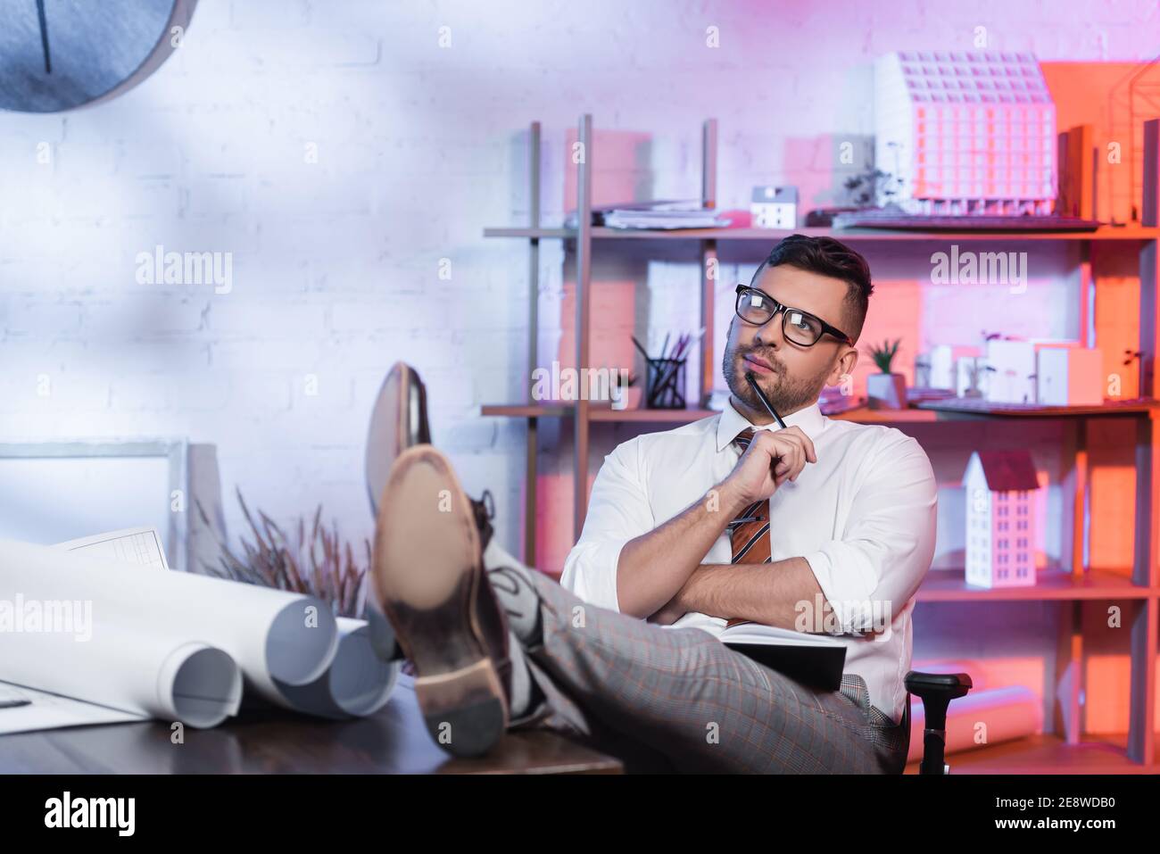thoughtful architect looking away while sitting with legs on desk Stock Photo