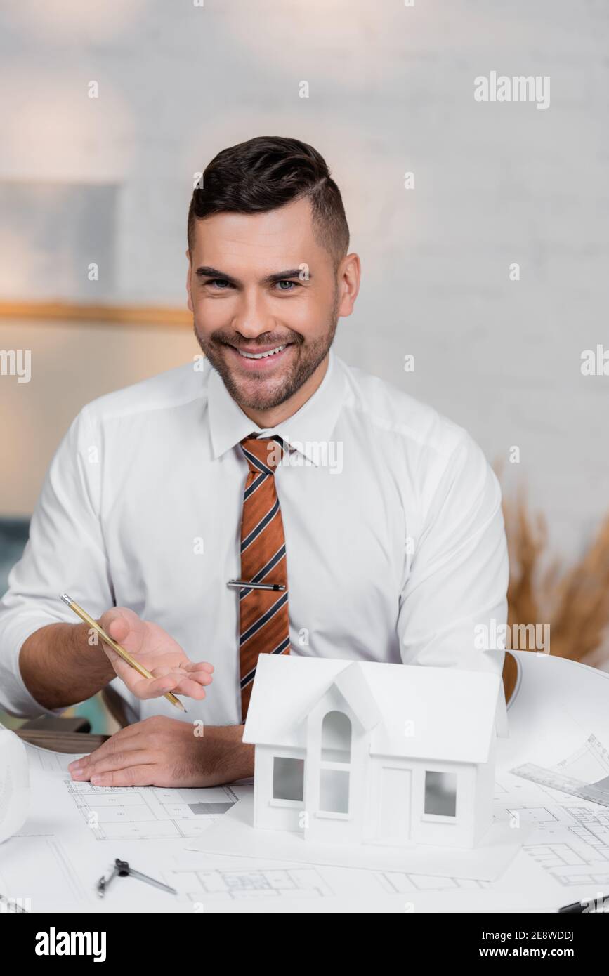 smiling architect looking at camera while pointing at house model Stock Photo