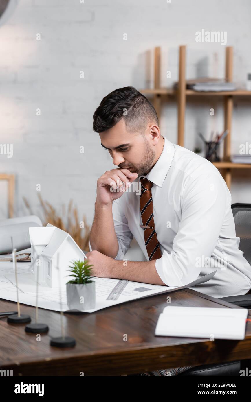 thoughtful architect sitting at workplace near house model and blueprint Stock Photo
