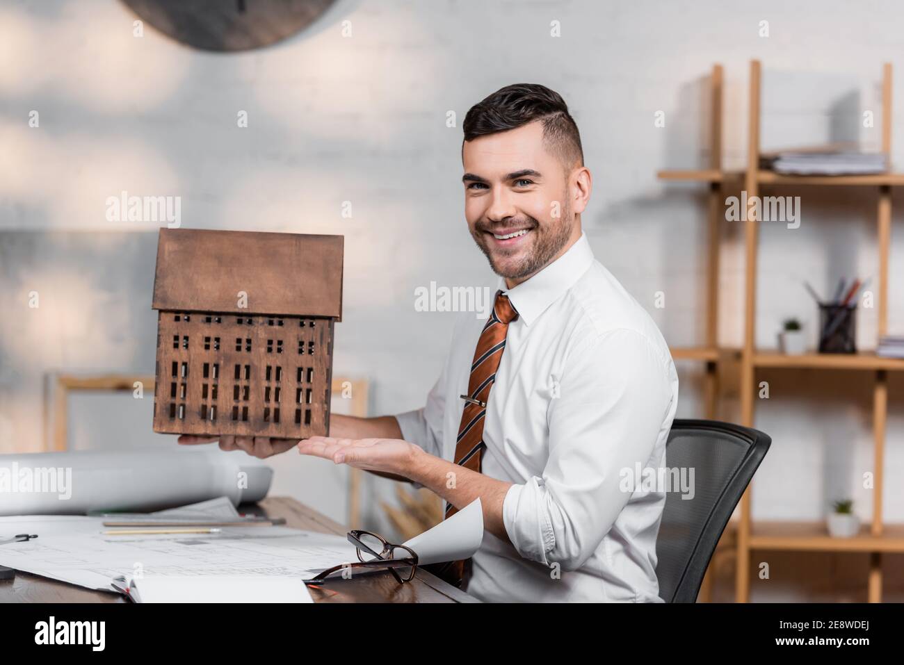 happy architect smiling at camera while pointing at house model at workplace Stock Photo