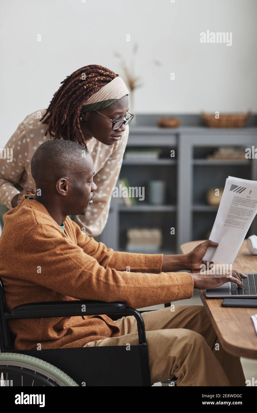 Vertical side view portrait of African-American man using wheelchair working from home with wife looking over his shoulder, copy space Stock Photo
