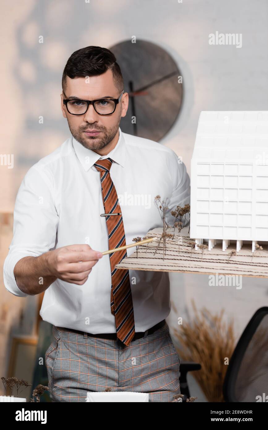 serious architect looking at camera while pointing with pencil at house model Stock Photo