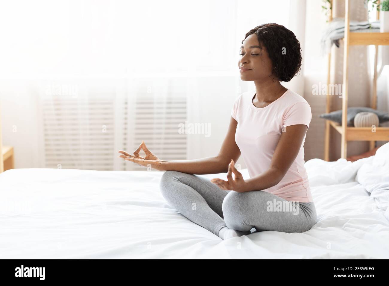Relaxed black woman sitting on bed and meditating Stock Photo