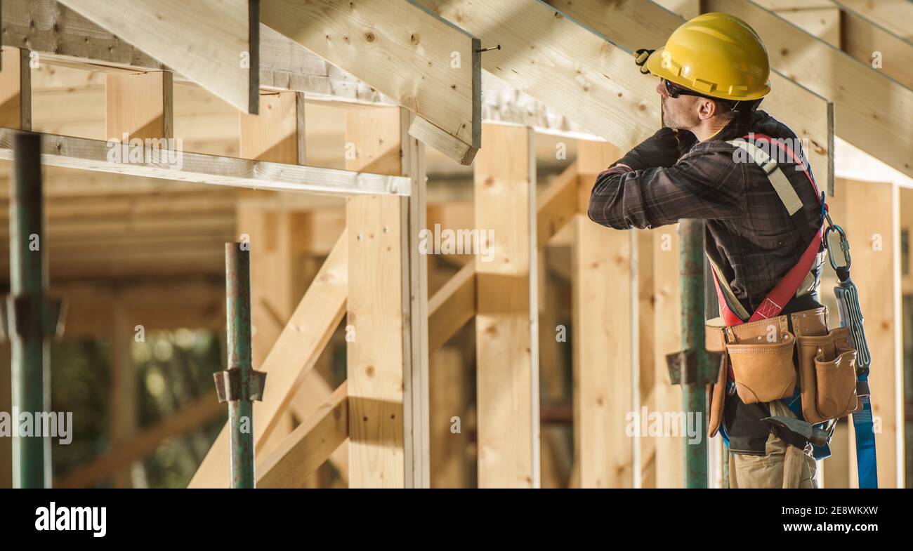 Caucasian Construction Worker in HIs 40s and Newly Built Wooden Roof Frame of the House. Construction Industry Theme. Stock Photo