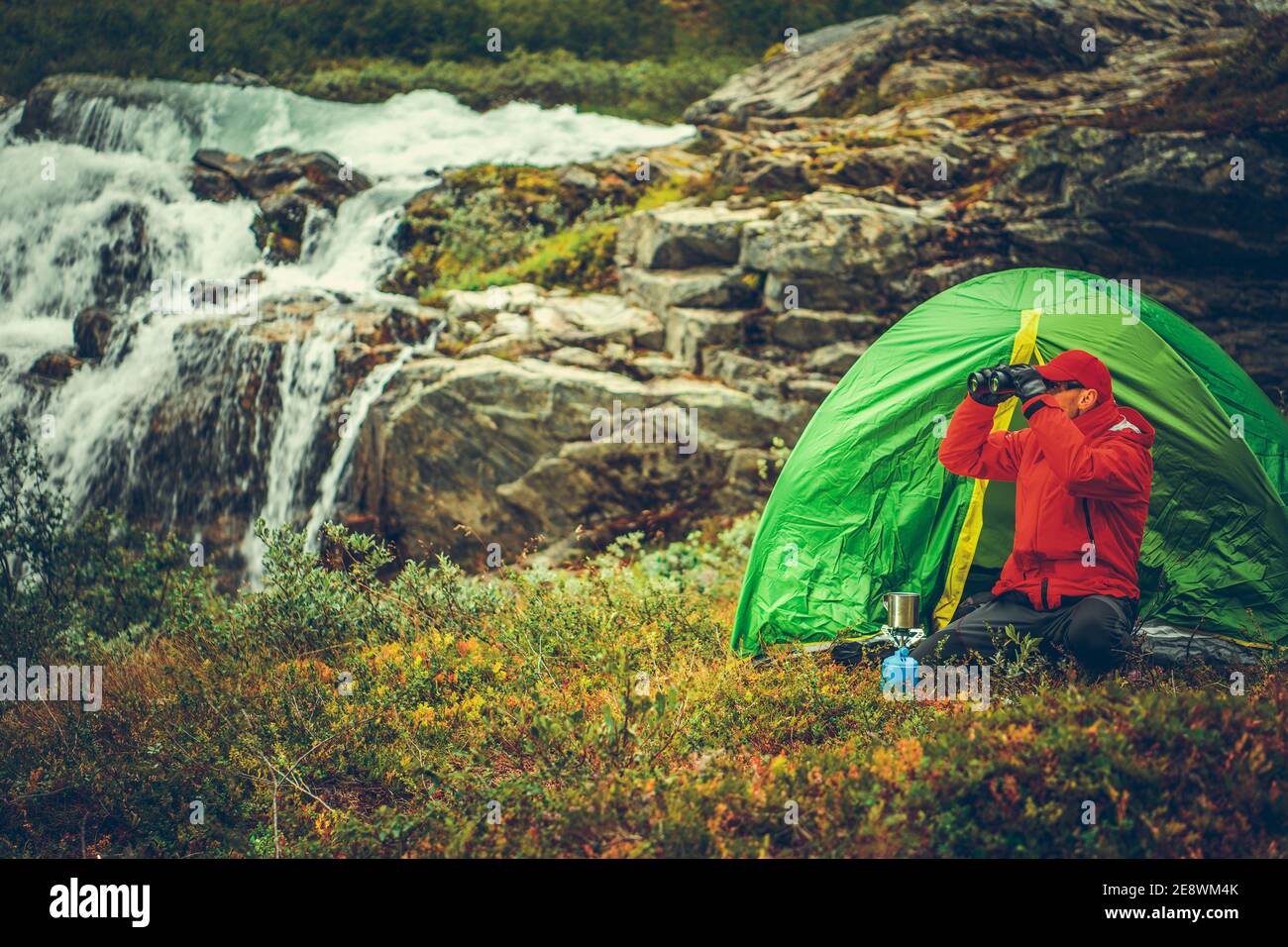 Caucasian Outdoor Men in Front of His Tent Spotting Something Using Binoculars. Scenic Landscape with Waterfall in Background. Wilderness Camping Spot Stock Photo