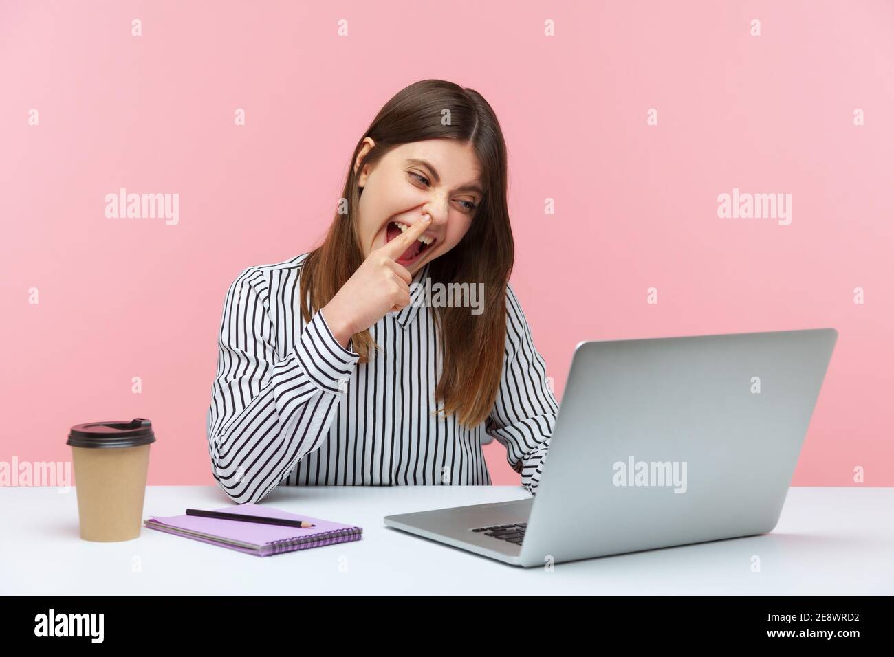 Lazy woman office worker having fun picking nose and looking into laptop screen with careless expression, talking on video call at workplace. Indoor s Stock Photo