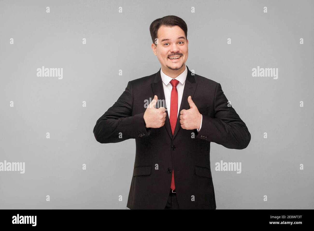 thumbs up handsome businessman in classic black suit showing like sign and looking at camera with toothy smile with mustache, laugh. Front view. Isola Stock Photo