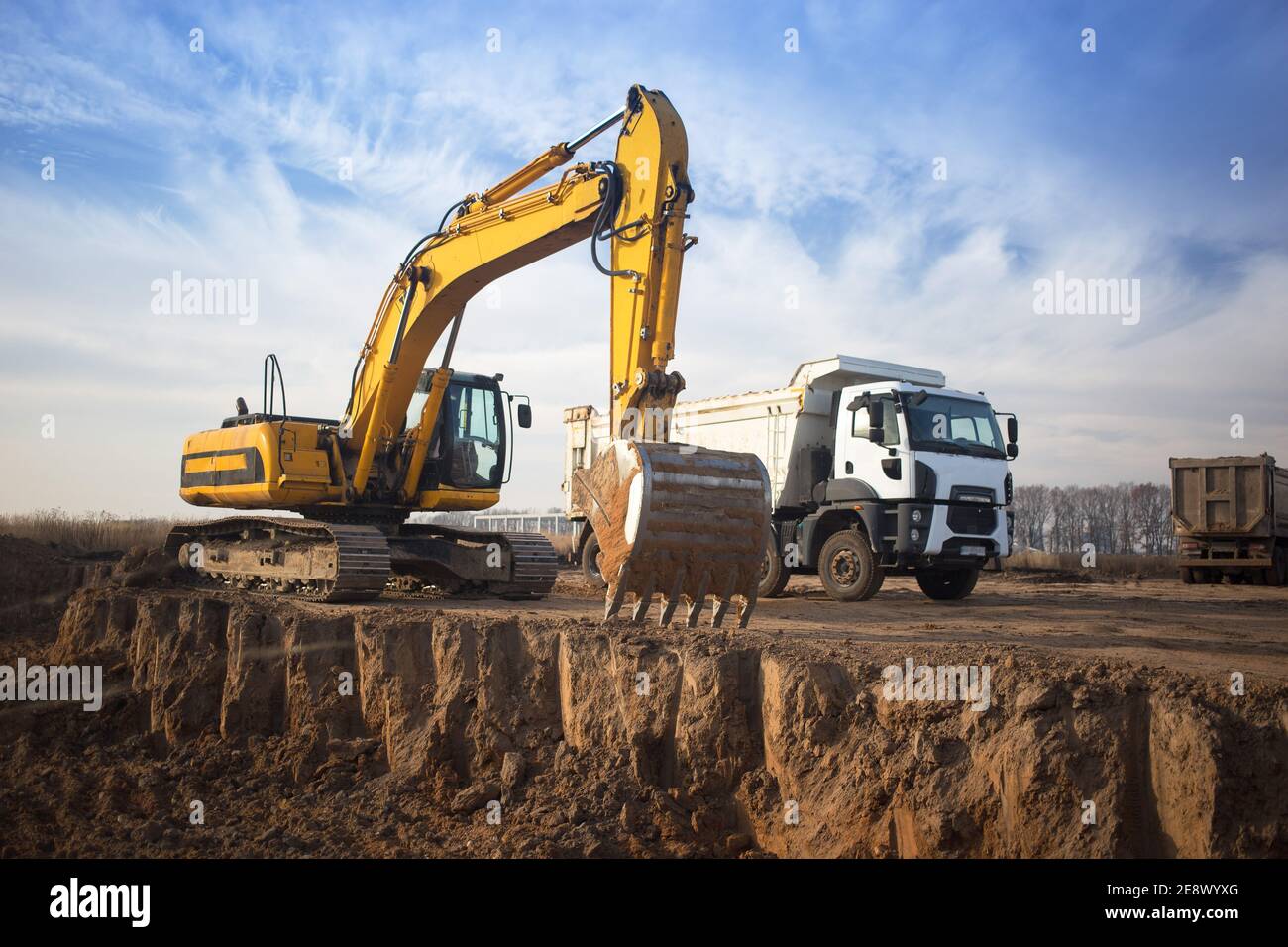 huge yellow crawler excavator and a construction dump truck standing next to it while working on a sunny day against a blue sky. Emphasis on the big b Stock Photo