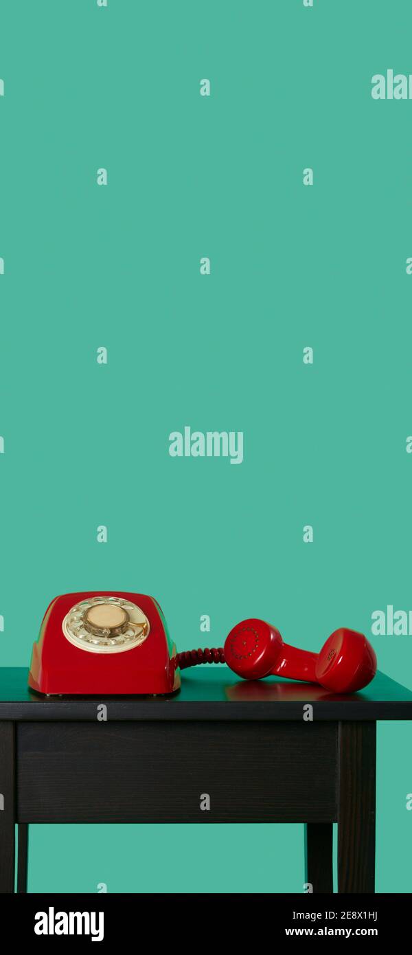 a red landline rotary dial telephone, with its handset off the hook on a black wooden table, on a green background, in a vertical format to use for mo Stock Photo