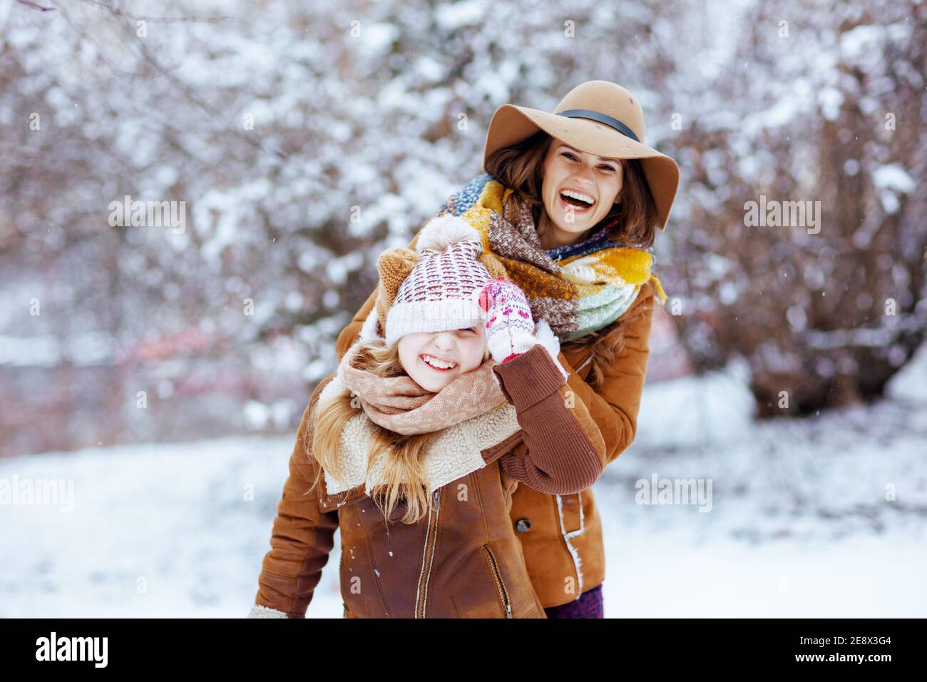 smiling modern mother and daughter in hats and sheepskin coats with mittens playing outdoors in the city park in winter. Stock Photo