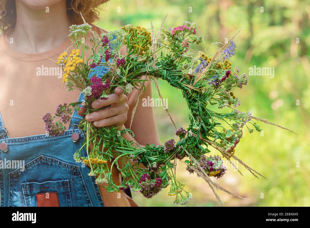 Woman in denim overalls holds a wreath of forest flowers. Close up. Forest background Stock Photo