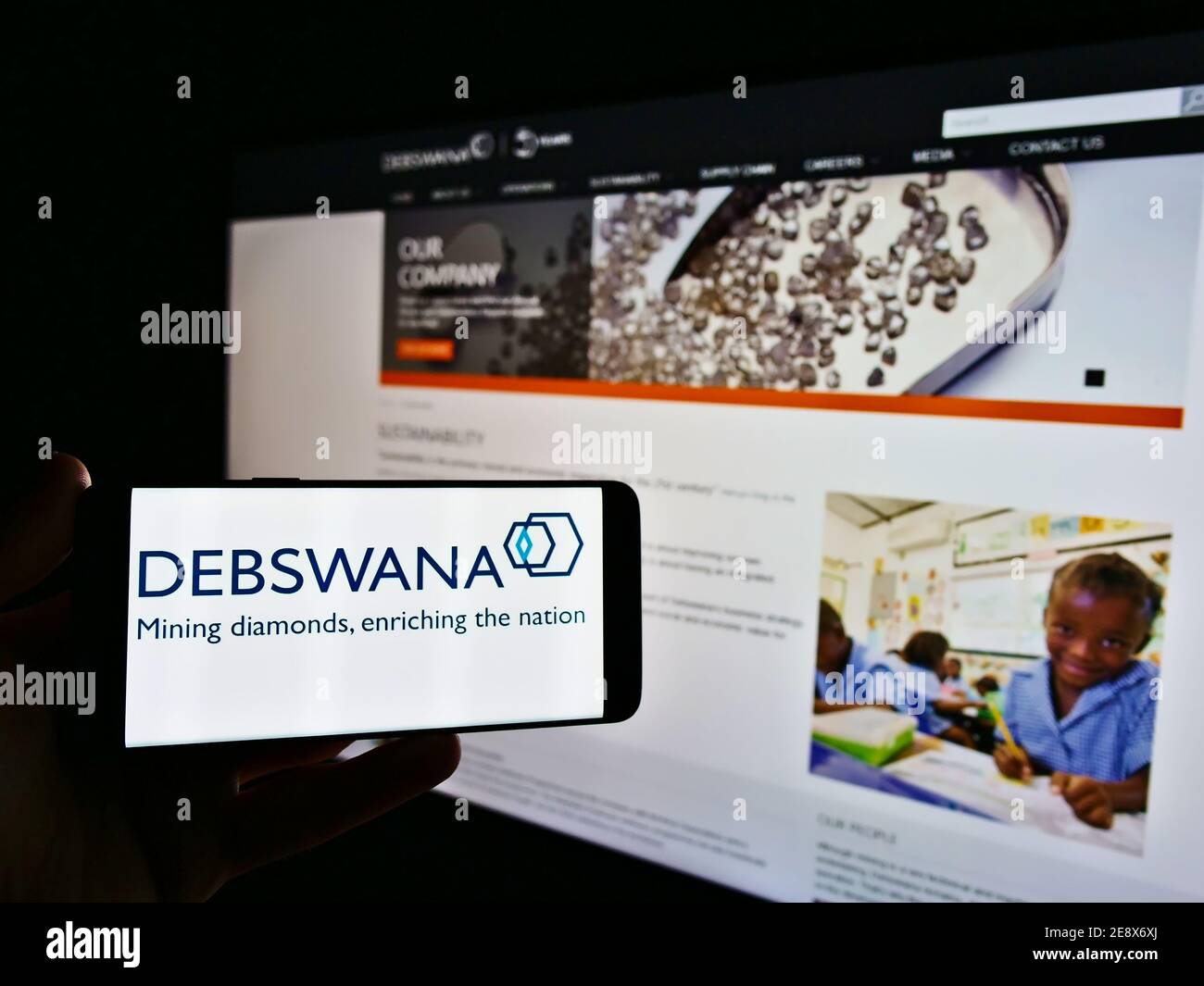 Person holding mobile phone with logo of mining company Debswana Diamond Company Limited on display in front of web page. Focus on phone screen. Stock Photo