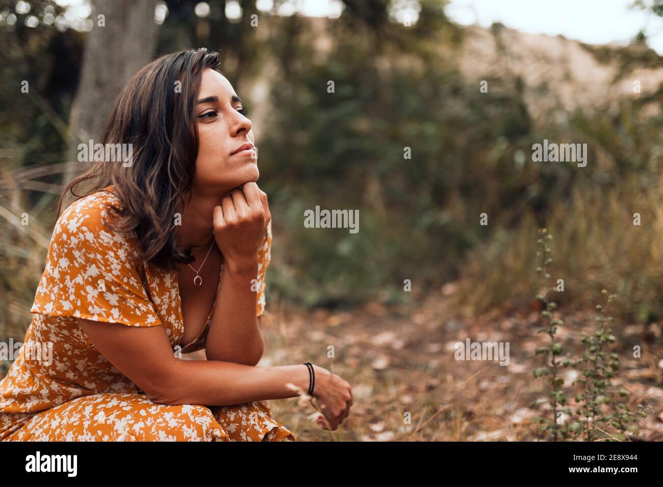 Thoughtful beautiful brunette woman wearing a dress squatting in the woods Stock Photo