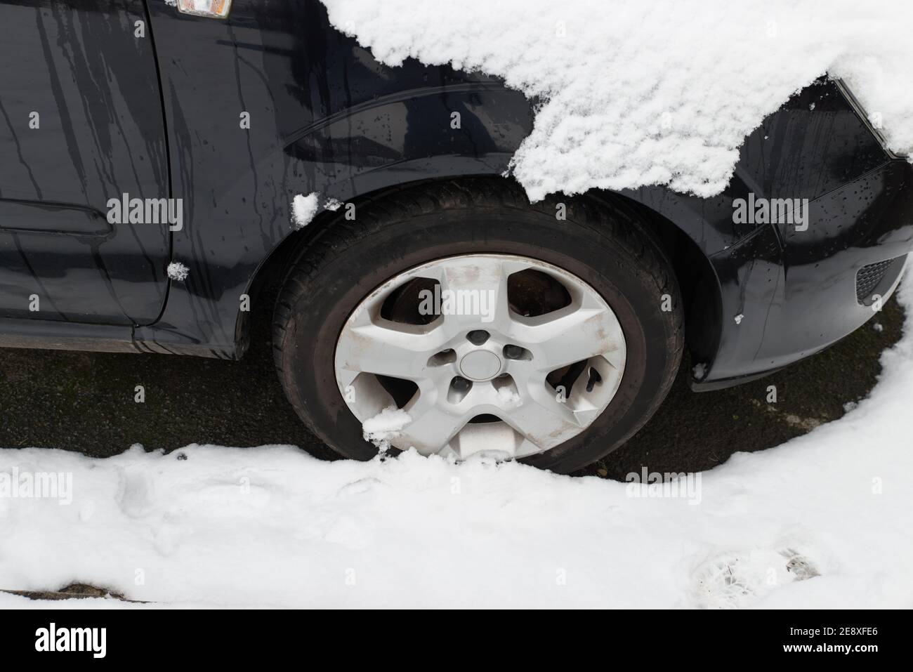 close up of black car wheel trim and rubber tyre  covered in snow.  The shiny car is parked up on a cold winters day Stock Photo