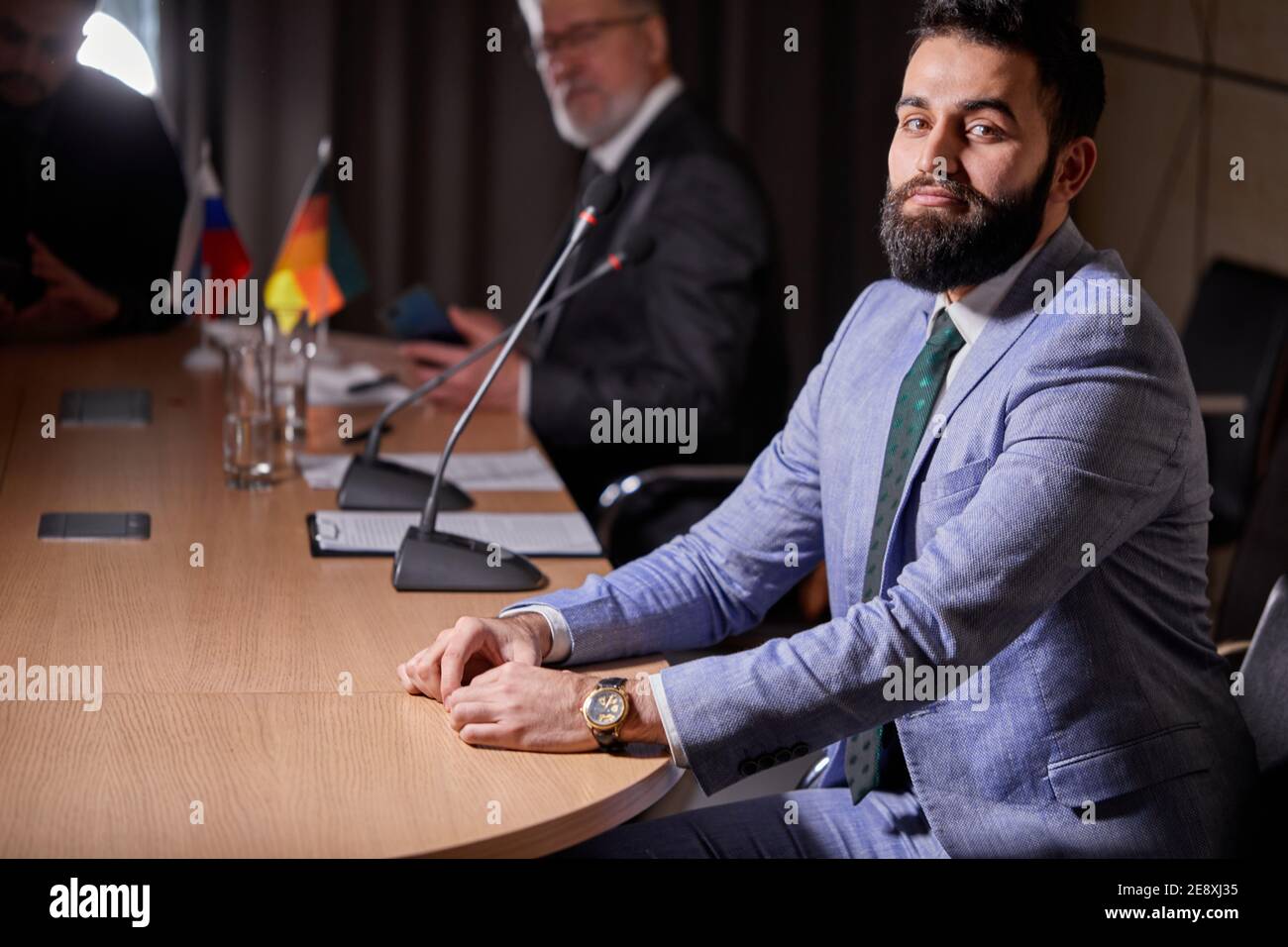 arabic male in suit sits at desk posing looking at camera, handsome smiling male executive satisfied with good job career posing in modern boardroom Stock Photo