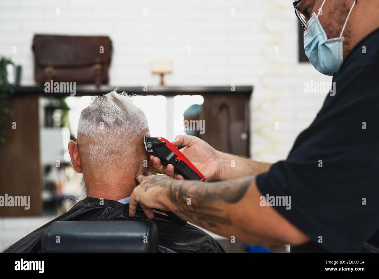 Male hairdresser cutting hair to hipster senior client while wearing face surgical mask - Young hairstylist working in barbershop during corona virus Stock Photo