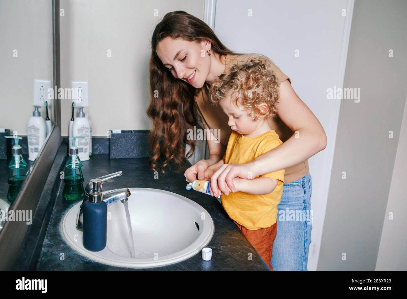 Young Caucasian mother helping boy toddler brush teeth in bathroom at home. Health hygiene and morning routine for children. Mom helping supporting te Stock Photo