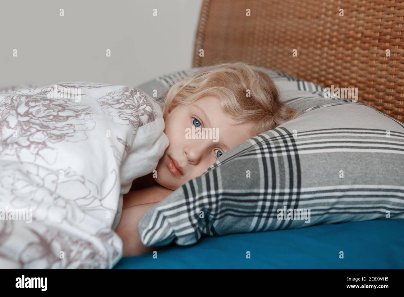 Pensive sad little Caucasian blonde child girl lying on pillow in bed at home. Child lying with open eyes thinking or dreaming. Candid authentic home Stock Photo