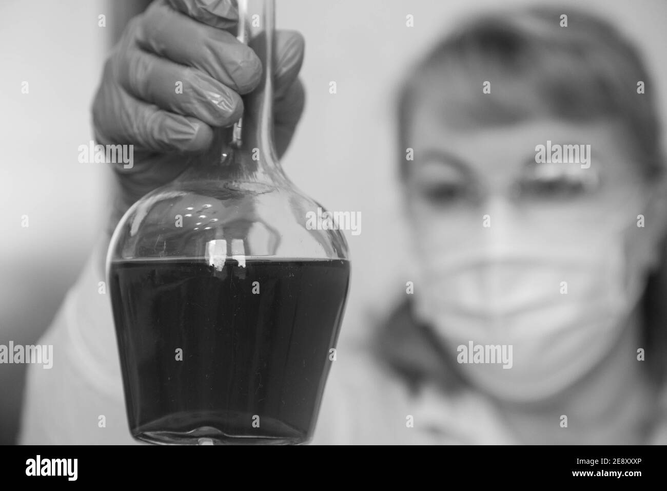 A woman in glasses and a mask is studying a vessel with liquid. Black and white Stock Photo