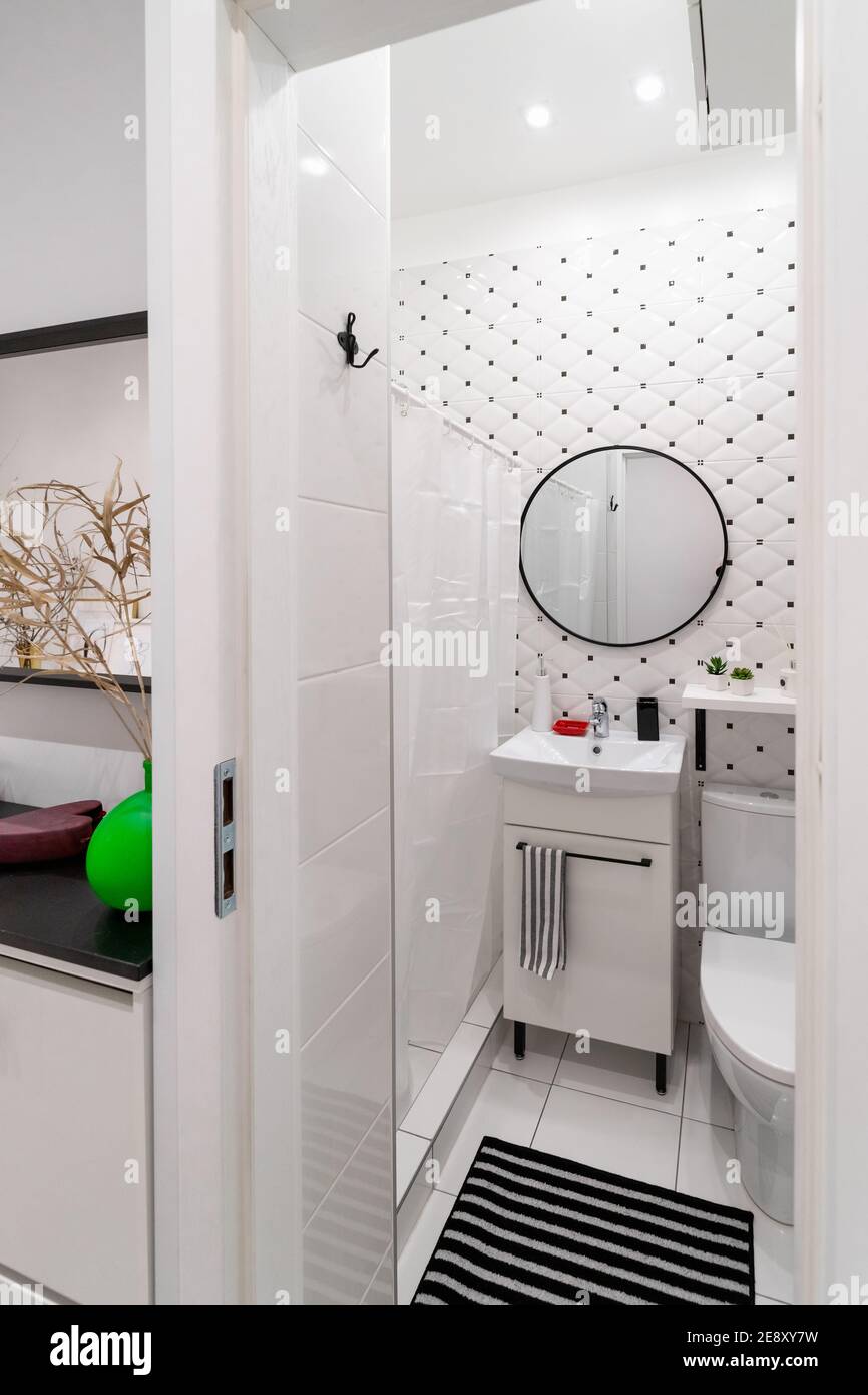 Modern tiled bright white-black bathroom and toilet in a small studio apartment. Interior view with cabinet and sink on it, round mirror, shower Stock Photo