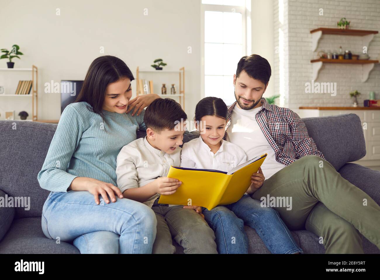 Parents with little daughter and son read fairy tales in a book sitting on the couch at home. Stock Photo