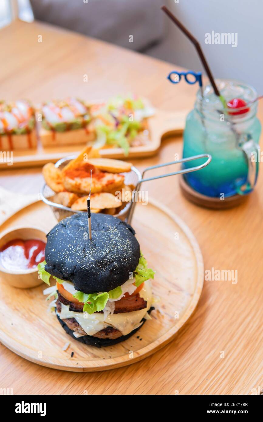 Black burger with meat patty, cheese, tomatoes, mayonnaise, french fries in a paper cup and glass of cold cola soda with ice. Dark wooden rustic table Stock Photo