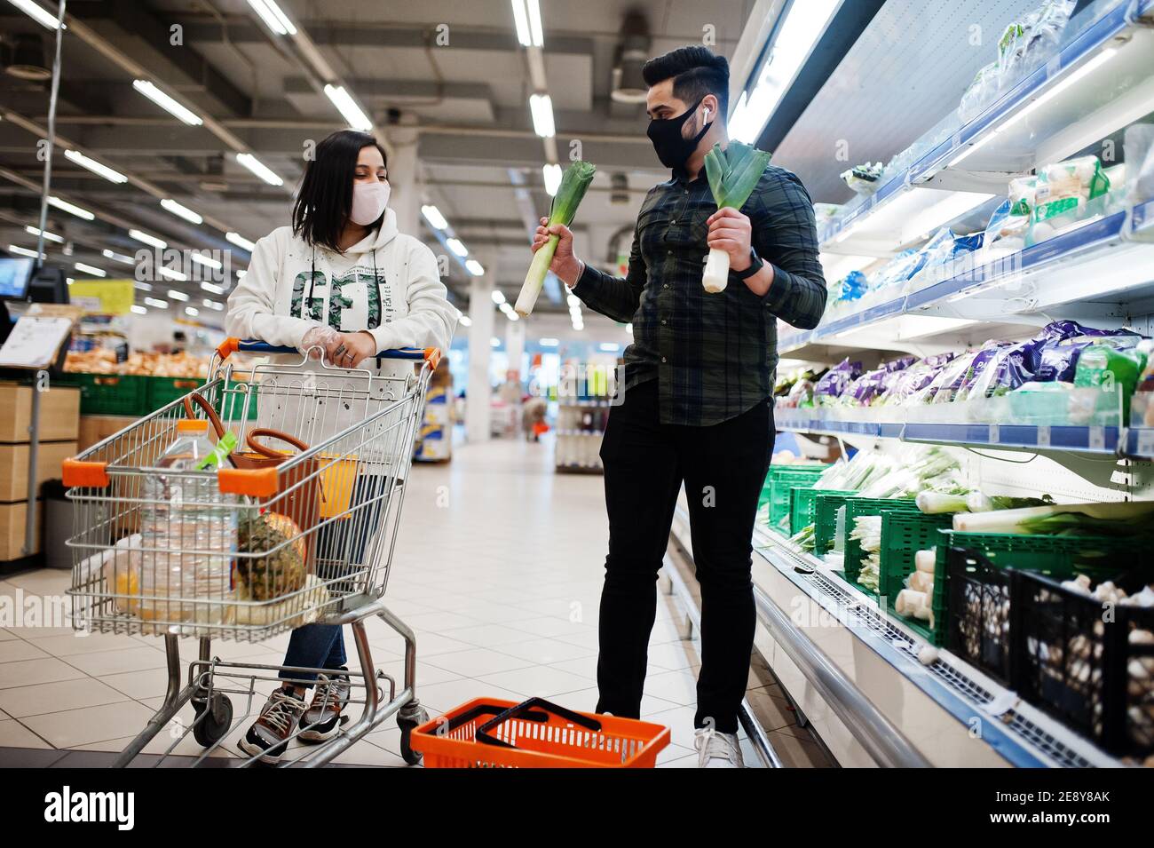 Asian couple wear in protective face mask shopping together in supermarket during pandemic. Taking vegetables from fridge. Stock Photo