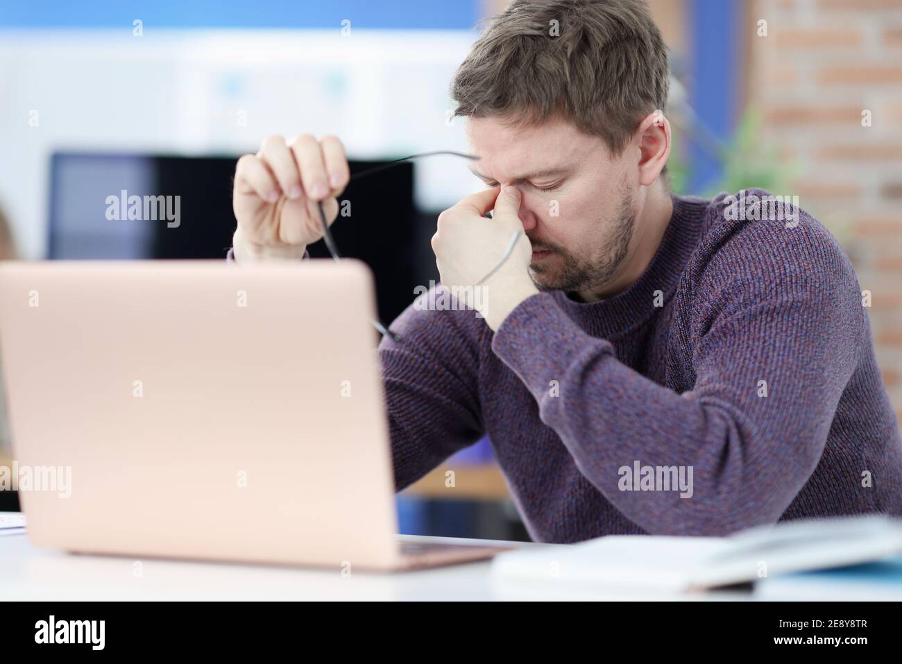 Tired man siting in front of laptop screen and holding glasses in hands ...