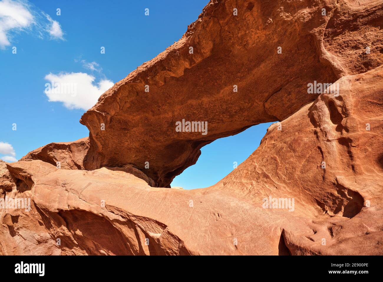Little arc or small rock window formation in Wadi Rum desert, bright sun shines on red dust and rocks, blue sky above Stock Photo