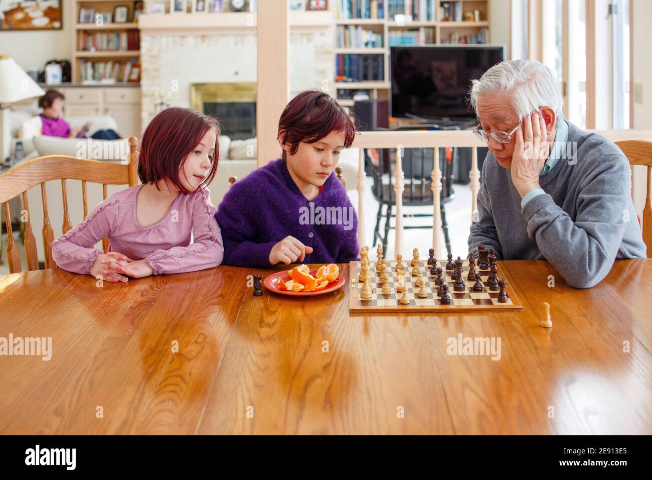 Two children play chess with grandfather and woman reads in background Stock Photo