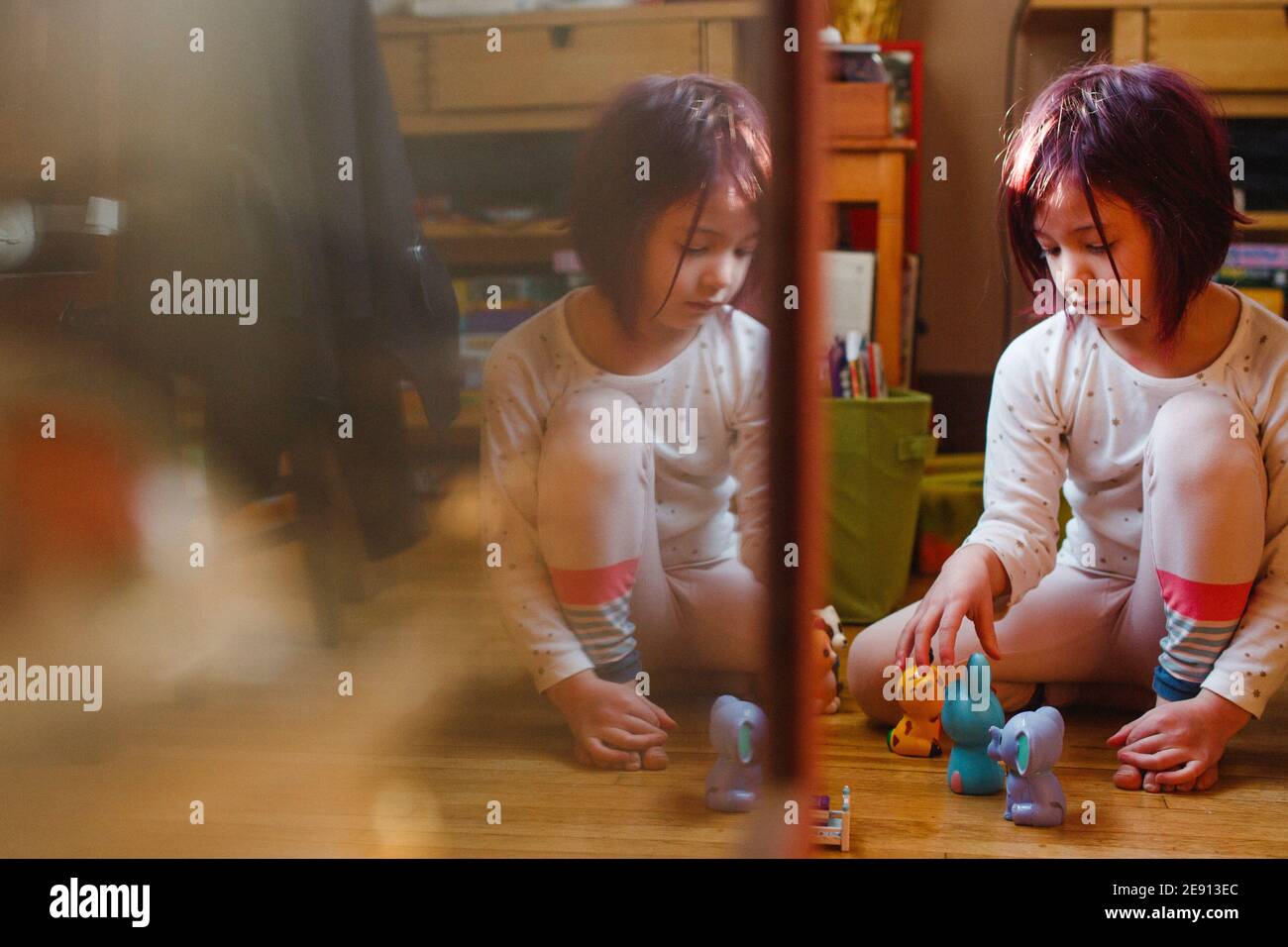A small child sits reflected in glass playing with toys on wood floor Stock Photo