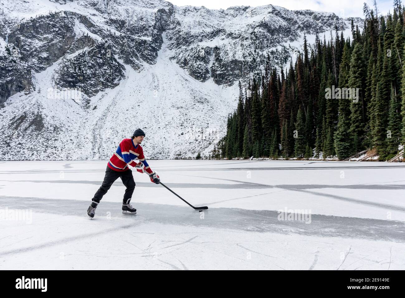 Young hockey player riding ice skates and manoeuvring puck with stick on frozen lake near snowy