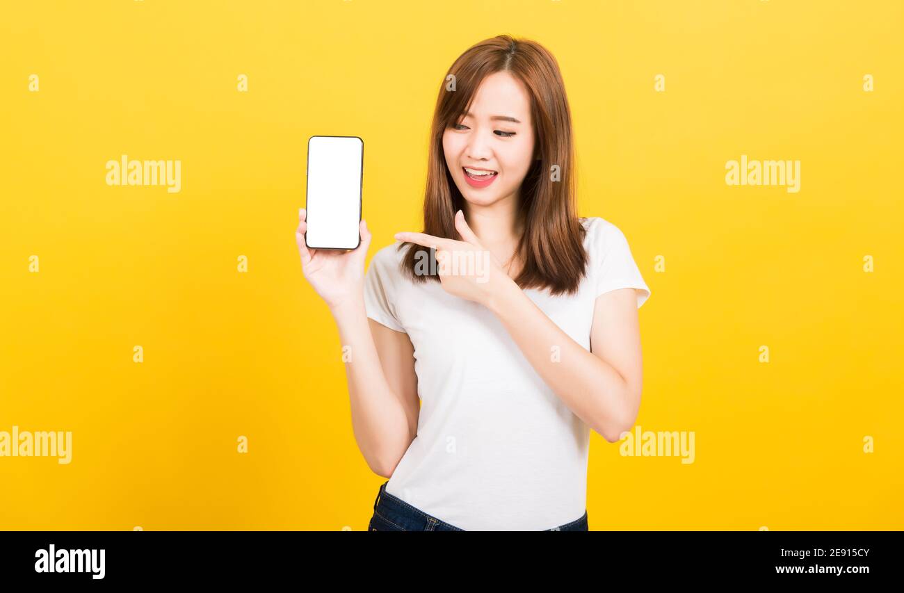 Asian happy portrait beautiful cute young woman smile standing wear t-shirt making finger pointing on smartphone blank screen looking to screen isolat Stock Photo