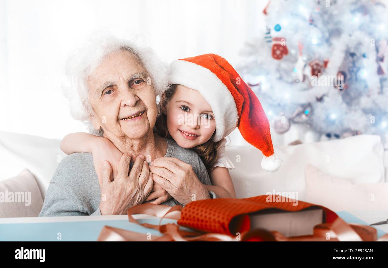 Smiling little granddaughter in santa hat sit hugging with grandma at Christmas Stock Photo