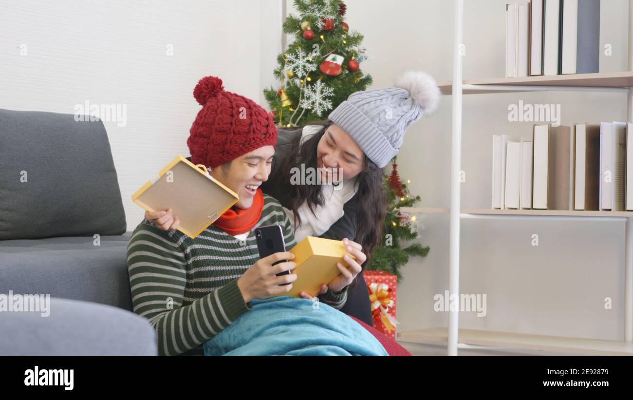 Happy Asian beautiful young family couple husband and wife in home living room, woman hide surprise giving gift box her boyfriend present during play Stock Photo