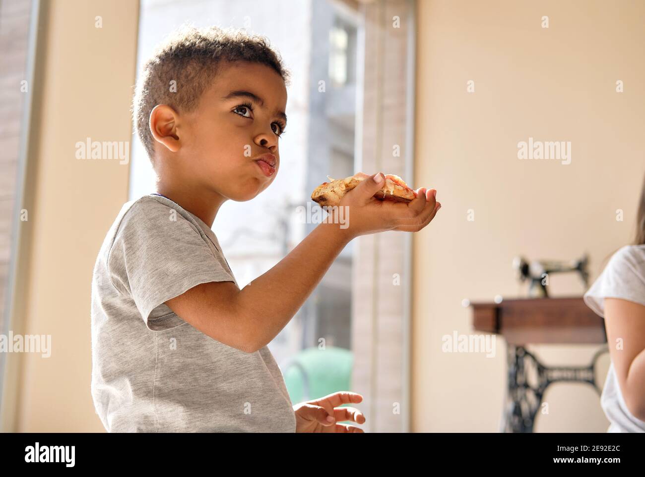 Three years old little mixed-race boy sit indoor eat pizza, enjoy fast food looking into distance. Italian cuisine small adorable lover, lunch time, l Stock Photo
