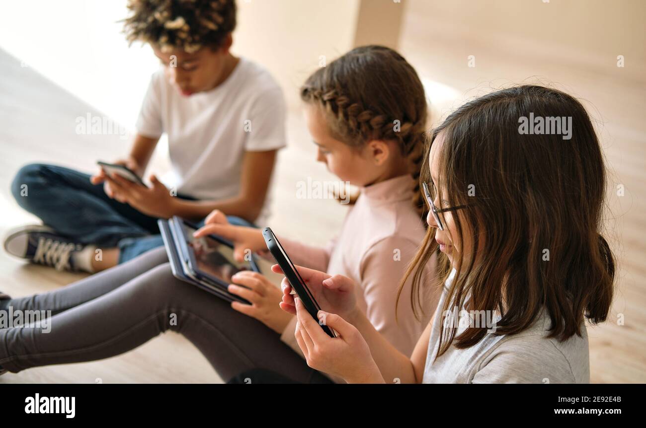 Three multi ethnic african, caucasian and korean little kids sit indoor using diverse wireless gadgets. Overuse of modern tech smart phones, digital d Stock Photo