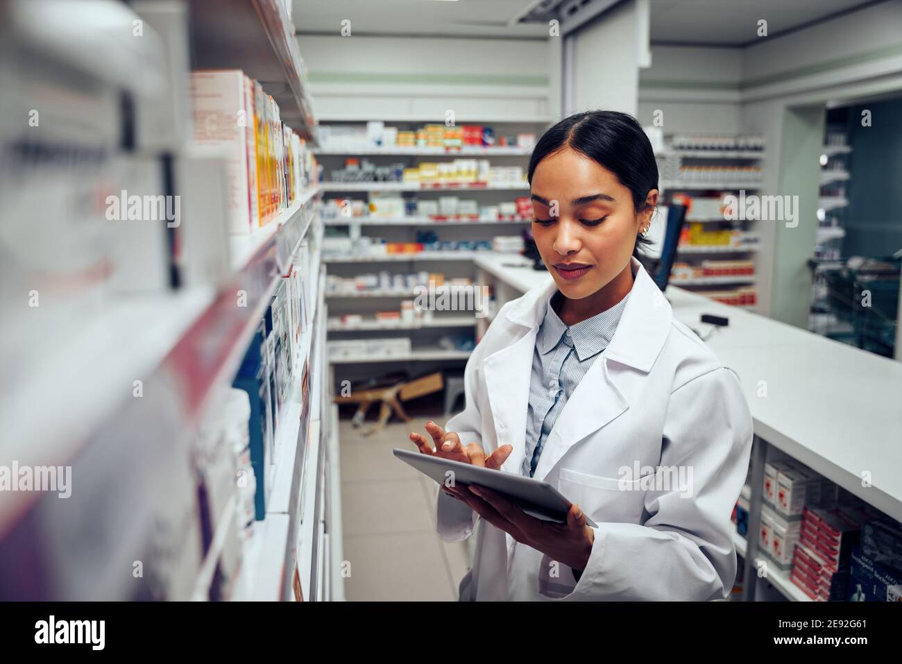 Young female pharmacist checking inventory of medicines in pharmacy using digital tablet wearing labcoat standing behind counter Stock Photo