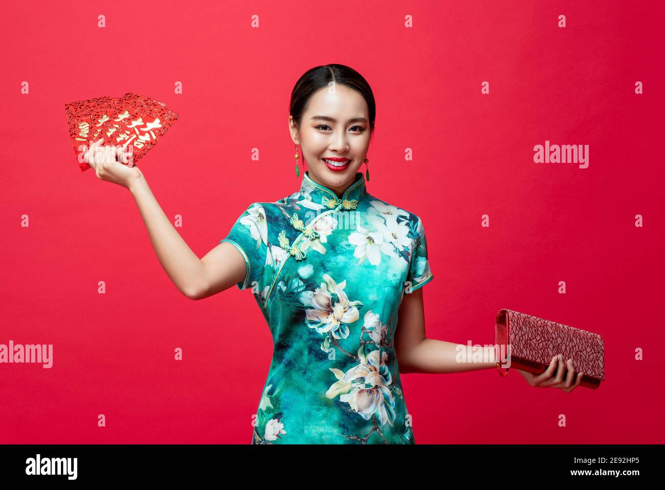 Smiling happy Asian woman wearing oriental qipao dress holding Ang Pao or red envelopes and wallet in hands on studio background for Chinese new year Stock Photo